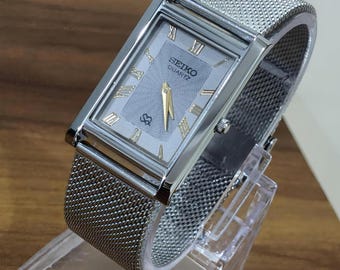 Könnte beinhalten: Eine rechteckige Seiko Quartz Armbanduhr mit silberfarbenem Zifferblatt, strukturiertem Zentrum, goldfarbenen Zeigern und goldfarbenen römischen Ziffern. Die Uhr hat ein poliertes silberfarbenes Metallgehäuse und ein silberfarbenes Milanaise-Armband.