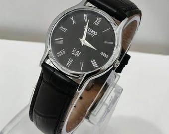 Könnte beinhalten: Eine Seiko Quartz Slim Armbanduhr mit rundem silberfarbenem Gehäuse, schwarzem Zifferblatt mit weißen römischen Ziffern und silberfarbenen Zeigern. Die Uhr ist mit einem schwarzen Lederarmband mit texturiertem Krokodilprägemuster ausgestattet.