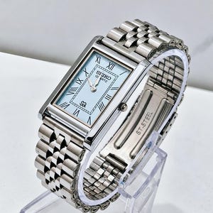 Pode incluir: Um relógio de pulso retangular Seiko Quartz com mostrador azul claro, ponteiros prateados e algarismos romanos pretos. O relógio possui uma pulseira de elos de metal prateado polido com um fecho gravado com o texto ST. STEEL.
