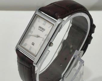 Könnte beinhalten: Eine rechteckige Seiko Quartz Armbanduhr mit silberfarbenem Gehäuse, weißem Zifferblatt mit schwarzen römischen Ziffern und einem dunkelbraunen Lederarmband mit Krokodilprägung sowie einer silberfarbenen Dornschließe.