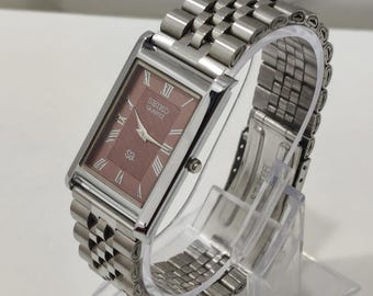 Könnte beinhalten: Eine rechteckige Seiko Quartz Armbanduhr mit einem strukturierten malvenfarbenen Zifferblatt, silberfarbenen römischen Ziffern und Zeigern. Die Uhr hat ein poliertes silberfarbenes Edelstahlgehäuse und ein mehrgliedriges Metallarmband.