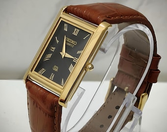 Könnte beinhalten: Eine rechteckige, goldfarbene Seiko Quartz Armbanduhr mit schwarzem Zifferblatt, goldenen römischen Ziffern und goldenen Zeigern. Die Uhr ist mit einem braunen Lederarmband mit geprägter Textur und einer goldfarbenen Schließe ausgestattet.