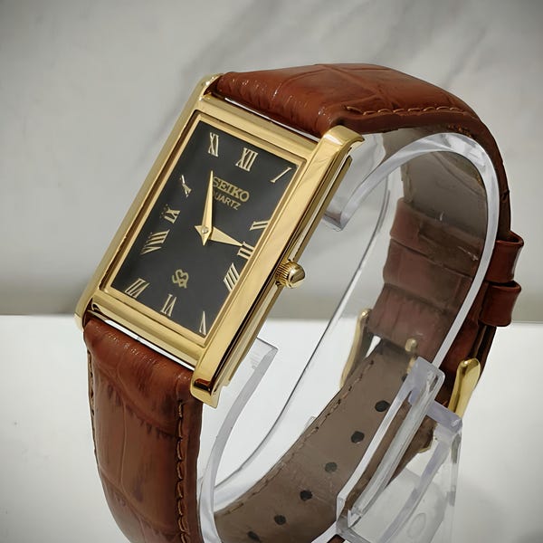May include: A rectangular gold-tone Seiko Quartz wristwatch featuring a black dial with gold Roman numeral hour markers and gold hands. The watch is paired with a brown leather strap that has a textured, embossed pattern and a gold-tone buckle.