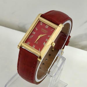 May include: A rectangular Seiko Quartz wristwatch featuring a gold-tone case, a deep red dial with gold-tone Roman numeral hour markers, and a textured red leather strap with a crocodile-embossed pattern and matching red stitching.