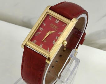 Könnte beinhalten: Eine rechteckige Seiko Quartz Armbanduhr mit goldfarbenem Gehäuse, einem tiefroten Zifferblatt mit goldfarbenen römischen Ziffern sowie einem strukturierten roten Lederarmband mit Krokodilprägung und passenden roten Nähten.