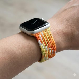 Puede incluir: Un reloj inteligente plateado con una correa de cordón trenzado con un patrón de chevrón repetitivo en tonos naranja, amarillo y blanco. La esfera del reloj es negra con manecillas blancas finas y la correa está hecha de cordón anudado.