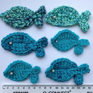 May include: Six crocheted fish appliques in various shades of blue and green, each featuring a small white bead for an eye. A white ruler is positioned at the bottom, showing the fish range from approximately 5 to 8 cm in length.