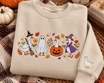 May include: A beige crewneck sweatshirt featuring an embroidered design of four geese in Halloween costumes, including a witch, a ghost, a pumpkin, and a wizard, surrounded by autumn flowers and leaves, with a small goose embroidered on the sleeve.