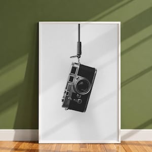 May include: A black and white framed wall art print featuring a vintage 35mm rangefinder camera hanging vertically by its black leather neck strap against a plain white background. The camera body is metallic silver and black with a lens.