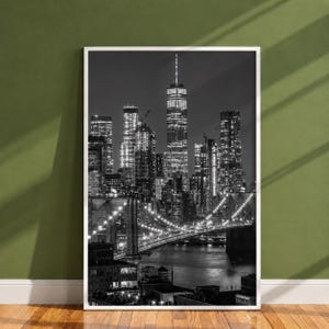 May include: A black and white framed wall art print featuring a nighttime cityscape of New York City. The image displays illuminated skyscrapers and the Brooklyn Bridge spanning across the water, captured in high contrast with detailed architectural lines.
