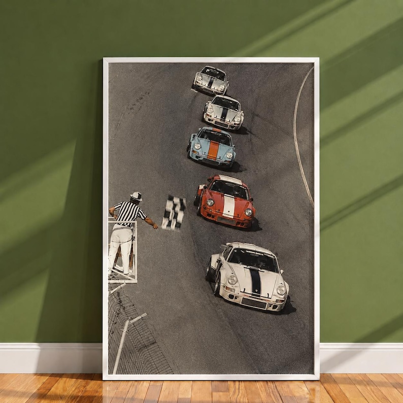 Pu&ograve; includere: Una stampa artistica da parete verticale incorniciata che mostra una vista dall'alto di cinque auto sportive d'epoca su una pista. Un commissario di gara in camicia a righe bianche e nere sventola una bandiera a scacchi al passaggio delle auto.