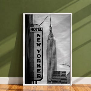 May include: A black and white framed wall art print featuring a vertical sign that reads HOTEL NEW YORKER in bold capital letters, positioned next to a detailed view of the Empire State Building against a cloudy sky in New York City.