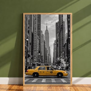 Puede incluir: Una fotografía enmarcada en blanco y negro de una calle de Nueva York con un taxi amarillo brillante en primer plano. El icónico Empire State Building se alza en el centro, enmarcado por altos rascacielos y arquitectura urbana.