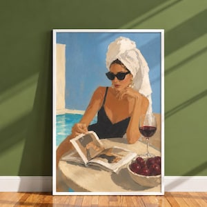 Op de afbeelding: Een ingelijste kunstdruk van een persoon in een zwart badpak met een witte handdoek op het hoofd en een zwarte zonnebril, zittend aan een tafel bij een blauw zwembad. De persoon leest een open boek naast een glas rode wijn en een schaal kersen.