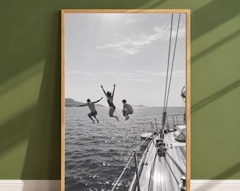 May include: A black and white framed wall art print depicting three individuals jumping into the ocean from the deck of a wooden sailboat. The scene captures the figures mid-air above the water, with the boat's rigging and deck visible in the foreground.
