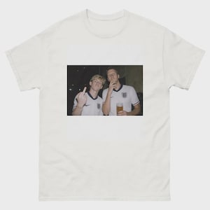 Könnte beinhalten: Ein weißes Kurzarm-T-Shirt mit einem rechteckigen Grafikdruck auf der Brust. Der Druck zeigt zwei Personen in weißen englischen Fußballtrikots, wobei eine Person ein Glas Bier hält und die andere Person den Mittelfinger in die Kamera zeigt.
