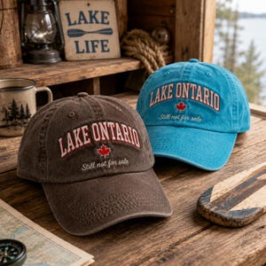 May include: Two baseball caps, one brown and one bright blue, each featuring the embroidered text LAKE ONTARIO in an arched font, a small red maple leaf icon, and the cursive script Still not for sale centered on the front panels.