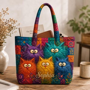 May include: A quilted tote bag featuring a colorful patchwork pattern and five fluffy, wide-eyed cats in purple, teal, orange, yellow, and blue. The name Sophia is printed in white script at the bottom center. The bag has two matching quilted handles.