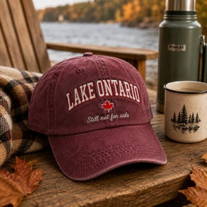 May include: A maroon baseball cap featuring the embroidered white text LAKE ONTARIO in an arched font, a small red maple leaf icon, and the cursive white text Still not for sale. The cap has a structured crown and a curved brim with stitching.