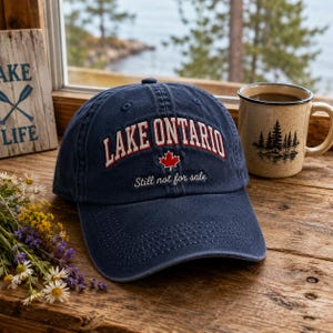 May include: A navy blue baseball cap featuring the embroidered text LAKE ONTARIO in red and white, a red maple leaf icon, and the white cursive text Still not for sale. The cap has a curved brim and a structured crown with stitched eyelets.