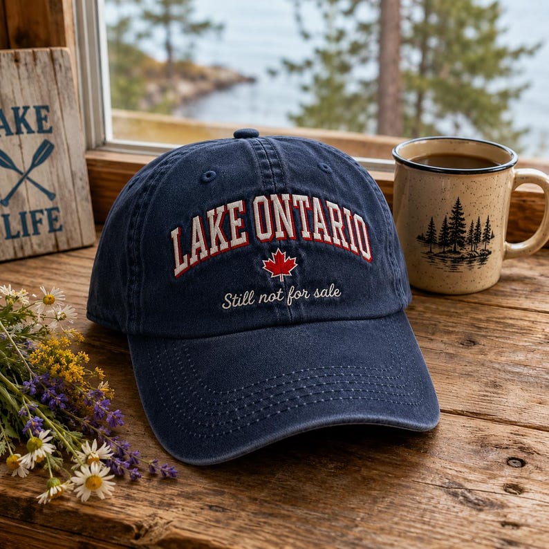 May include: A navy blue baseball cap featuring the embroidered text LAKE ONTARIO in red and white, a red maple leaf icon, and the white cursive text Still not for sale. The cap has a curved brim and a structured crown with stitched eyelets.