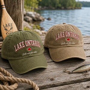 May include: Two baseball caps in olive green and tan, each featuring the embroidered text LAKE ONTARIO in red, a small red maple leaf icon, and the white cursive text Still not for sale centered on the front panels of the cotton fabric.