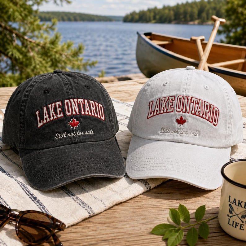 May include: Two baseball caps, one charcoal black and one white, each featuring the embroidered red text LAKE ONTARIO in an arched font, a small red maple leaf icon, and the cursive white text Still not for sale on the front panels.