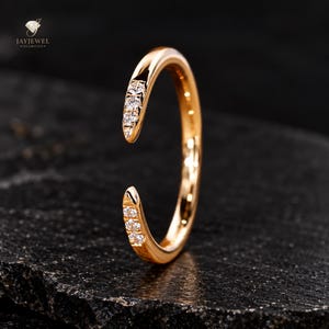 May include: An open-style rose gold ring featuring a polished band with three small, round, clear stones set into each of the two tapered, open ends. The ring is positioned vertically, showcasing its smooth, reflective metal surface and design.