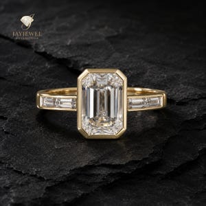 May include: A yellow gold ring featuring a large emerald-cut center diamond in a bezel setting, flanked by two smaller baguette-cut diamonds on each side of the band. The ring is marked with the JAYJEWEL DIAMOND logo in the upper left corner.