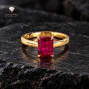 Pode incluir: Um anel de ouro com uma pedra preciosa vermelha vibrante de corte esmeralda retangular presa por quatro garras. A banda é de ouro liso e polido. O logótipo JAYJEWEL DIAMOND é visível no canto superior esquerdo com um ícone de diamante.