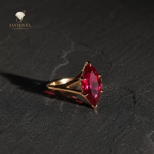 May include: A gold ring featuring a marquise-cut red gemstone held by a prong setting. The band has a split design that connects to the base of the stone. The text JAYJEWEL DIAMOND is visible in the upper left corner of the image.