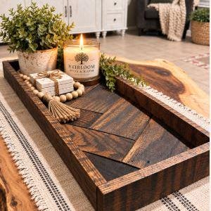 May include: A rectangular wooden tray with a dark brown chevron pattern base. Inside the tray sits a potted green plant in a speckled white pot, a lit candle in a glass jar labeled Heirloom Home & Bloom, and a stack of white coasters with a tassel.