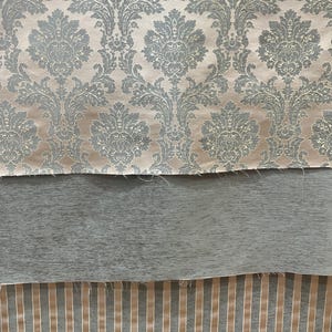 May include: Three stacked fabric swatches. The top layer features a light beige background with a repeating grey damask pattern. The middle layer is a solid, textured grey fabric. The bottom layer displays a vertical stripe pattern in grey and beige.
