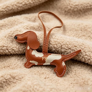 May include: A brown and white dachshund-shaped leather ornament featuring a loop for hanging. The dog figure has white patches on its body, a black nose, and the letters FS embossed in gold on its side, with visible white stitching along the edges.