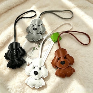 May include: Four dog-shaped bag charms made of textured material in black, grey, white, and brown. Each charm features a long strap for attachment, a rounded head with floppy ears, and a small snout. The charms are arranged in a cluster.
