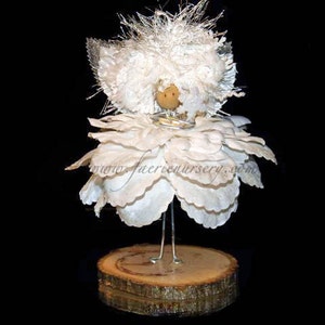 May include: A white fairy figurine with a white flower-shaped skirt and a wooden base. The fairy has a white feathery halo and a wooden head with a painted face.