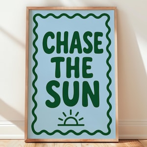 Op de afbeelding: Een rechthoekige wandprint in een lichthouten lijst met een lichtblauwe achtergrond en een donkergroene golvende rand. De tekst CHASE THE SUN staat in donkergroene hoofdletters boven een klein donkergroen zonicoon onderaan.