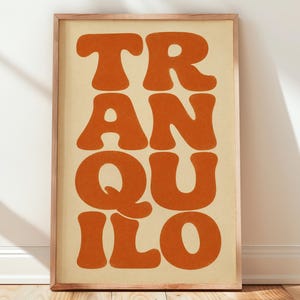 May include: A rectangular wall art print in a light wood frame featuring the word TRANQUILO in large, bold, burnt orange letters stacked in two columns against a solid, light beige background. The typography has rounded, soft edges.
