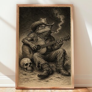 May include: A framed wall art print featuring a detailed black and white illustration of a toad wearing a cowboy hat and boots, smoking a cigar, and playing an acoustic guitar. A human skull rests on the ground beside the seated, guitar-playing toad.