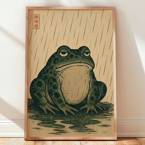 May include: A framed vertical print featuring a dark green, spotted toad sitting in a puddle during a rainstorm. The background is a light tan color with vertical lines representing rain. A red square seal with Japanese characters is in the top left.