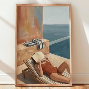 May include: A framed art print depicting a person in a rust-colored swimsuit lying on a wooden lounge chair by a pool, reading a book. A blue and white striped towel rests on a stone ledge nearby, with a view of the sea in the background.