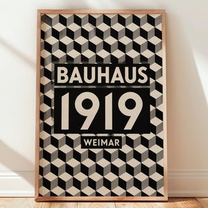 May include: A rectangular framed poster featuring a repeating geometric pattern of black, white, and gray cubes. Centered on the print is a black horizontal block containing the white text BAUHAUS, 1919, and WEIMAR in a bold sans-serif font.