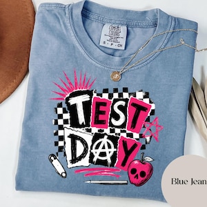 Puede incluir: Una camiseta azul de manga corta con un estampado gráfico que incluye el texto "TEST DAY". El diseño presenta un patrón de cuadros en blanco y negro, una estrella rosa, un imperdible, un lápiz y una manzana roja con cara de calavera.