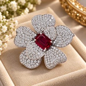 May include: A flower-shaped brooch featuring five petals densely encrusted with small, clear, sparkling stones. At the center of the floral design sits a single, rectangular, deep red gemstone with faceted edges, set against a metallic frame.