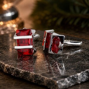 May include: A pair of silver-toned cufflinks featuring rectangular, deep red faceted stones held within a polished metal frame. The design includes a hinged toggle mechanism on the back of each piece, showcasing a clean, metallic finish.