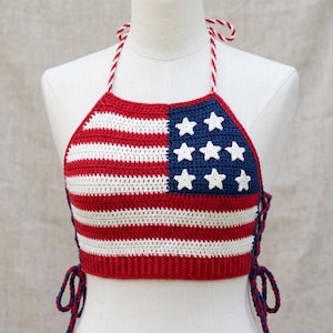 May include: A crochet halter top featuring a red, white, and blue American flag pattern. The garment includes a blue square section with eight white stars, horizontal red and white stripes, red and white braided neck ties, and blue side laces.