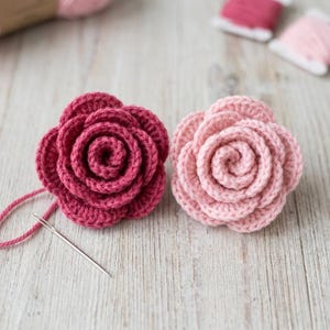 Könnte beinhalten: Zwei handgefertigte Häkelblumen, eine in tiefem Magenta und eine in zartem Pastellrosa, mit geschichteten Blütenblättern in Spiralform. Eine einzelne silberne Nähnadel liegt neben der magentafarbenen Blume mit einem Stück passendem Garn.