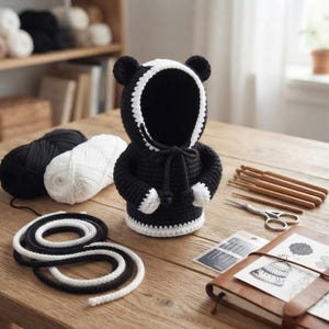 Può includere: Una felpa con cappuccio all'uncinetto bianca e nera a forma di orso, dotata di cappuccio, coulisse e orecchie arrotondate. Il capo presenta un corpo nero tinta unita con bordi bianchi lungo l'apertura del cappuccio, i polsini e l'orlo.