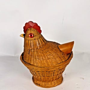 May include: A woven wicker basket shaped like a chicken with a light brown natural finish. The chicken features a bright red painted wooden comb on top of its head, a small yellow beak, and a single round red eye on the side of its face.