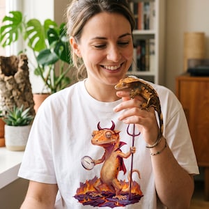 May include: A white t-shirt featuring a graphic of a gecko dressed as a devil with horns, a cape, and a trident, standing on lava. The text below the graphic reads L'ENFER COMMENCE A 30°C. The design includes a compass held by the gecko.
